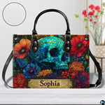 Stained glass skull handbag on white wooden table beside floral vase with texture swatch inset.