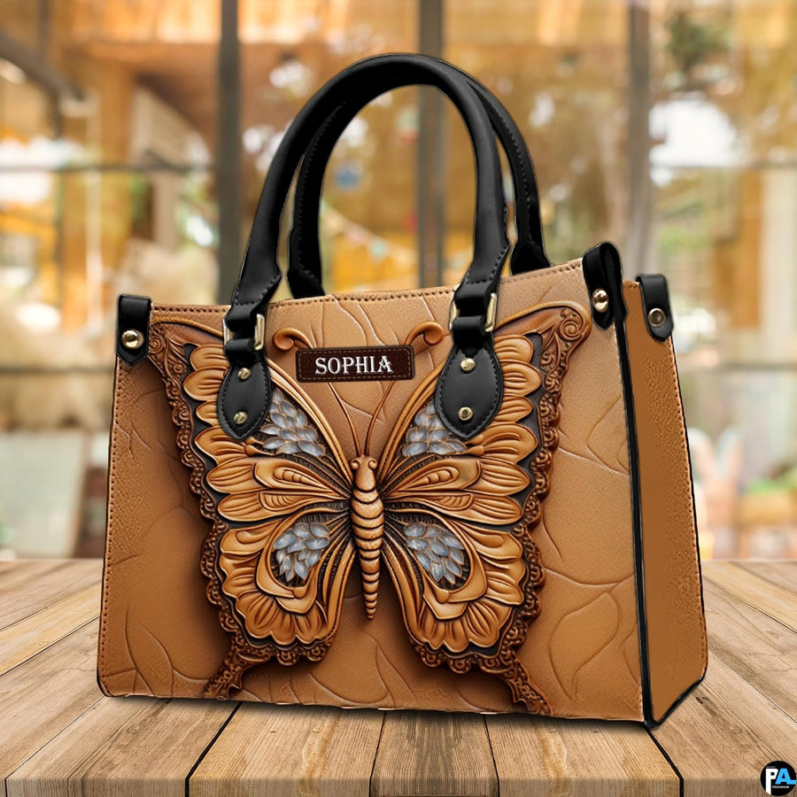 Sculpted tan butterfly handbag resting on a sunny outdoor wooden deck in front of large glass windows.