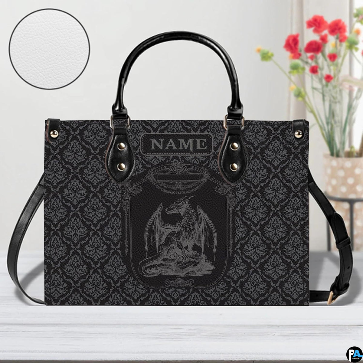 Dragon crest handbag on white wooden table beside floral vase with texture swatch inset.