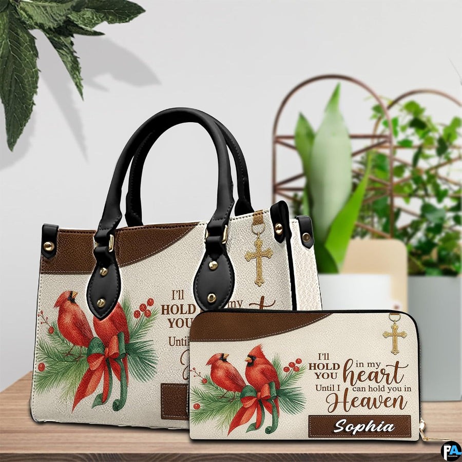 Red cardinal remembrance handbag and matching wallet displayed on a wooden surface beside green foliage.