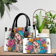 Praying Madonna handbag and coordinating wallet displayed on a wooden table beside green potted plants.