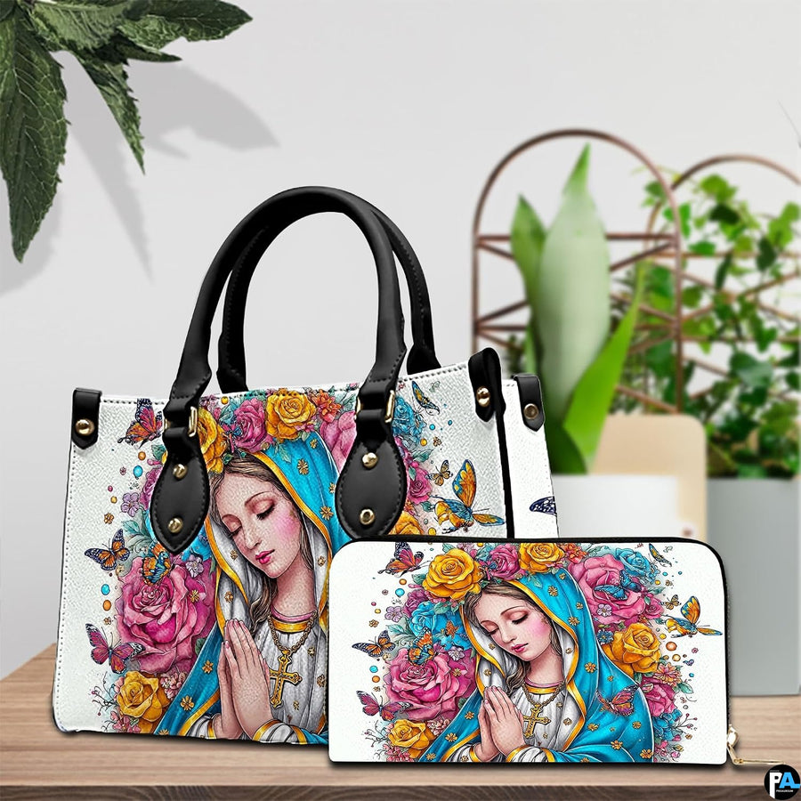 Praying Madonna handbag and coordinating wallet displayed on a wooden table beside green potted plants.