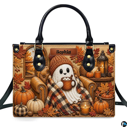 Front view of autumn satchel with embossed ghost in beanie sipping cocoa in armchair with pumpkins and candles.