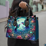 Model holding handbag outdoors showcasing twin hummingbirds and glowing moonlit botanical artwork.