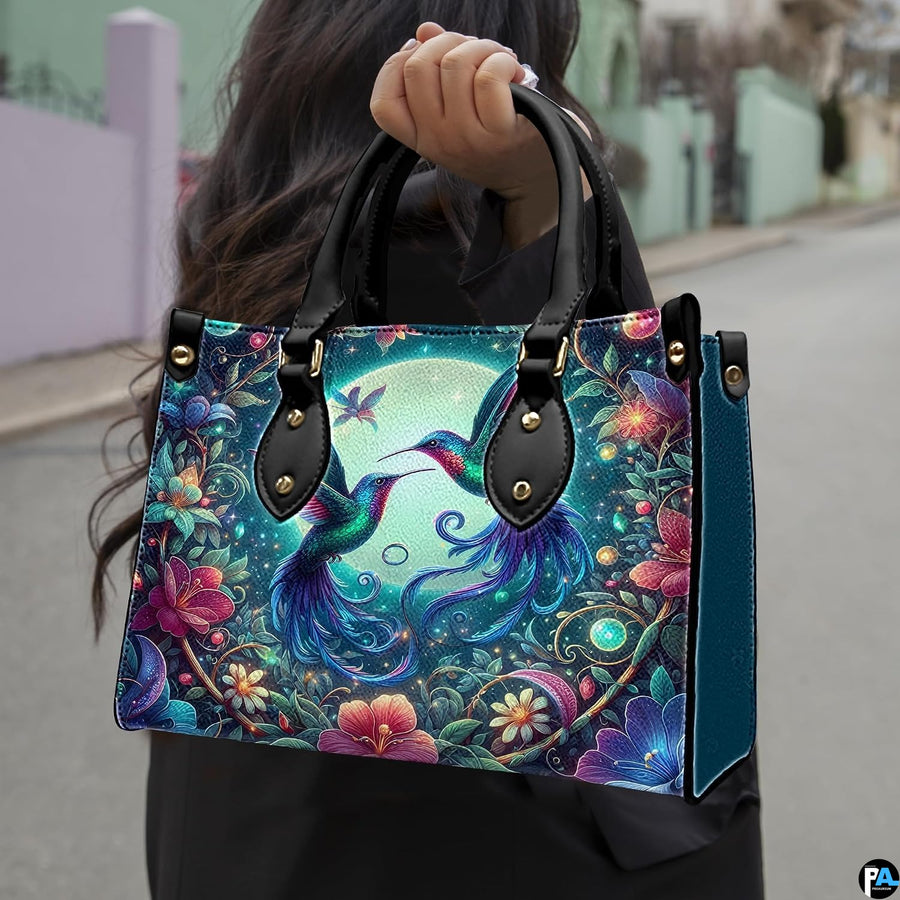 Model holding handbag outdoors showcasing twin hummingbirds and glowing moonlit botanical artwork.