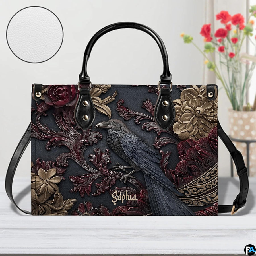 Gothic raven handbag displayed on white table beside floral bouquet.