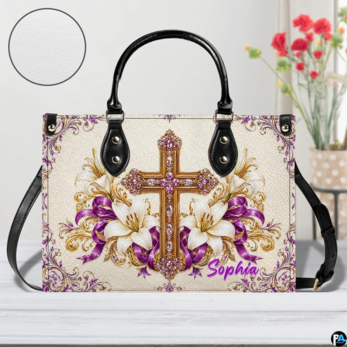 Top-handle handbag with purple cross and white lily artwork resting on a white wooden surface.