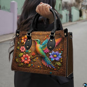 Model holding bronze handbag outdoors displaying colorful hummingbird and floral branch artwork.