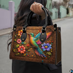 Model holding bronze handbag outdoors displaying colorful hummingbird and floral branch artwork.