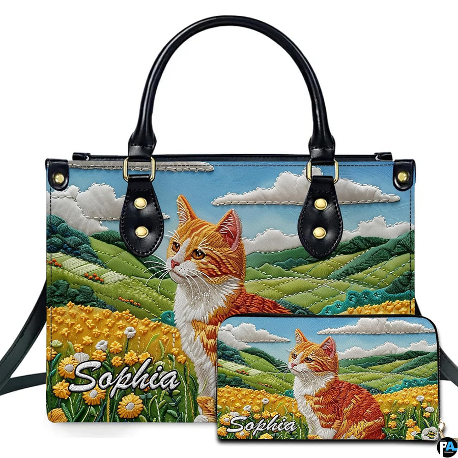 Embroidered ginger tabby cat seated in a yellow buttercup meadow beside rolling green hills.