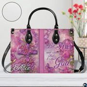 Front view of pink handbag featuring wooden cross draped with blooming lilies and flying butterflies.