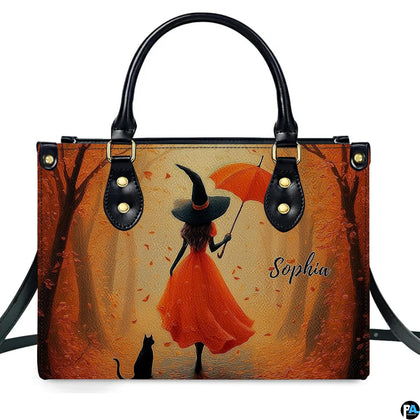 Autumn witch in orange gown with umbrella and black cat walking down golden tree path.