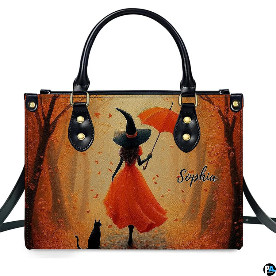 Autumn witch in orange gown with umbrella and black cat walking down golden tree path.