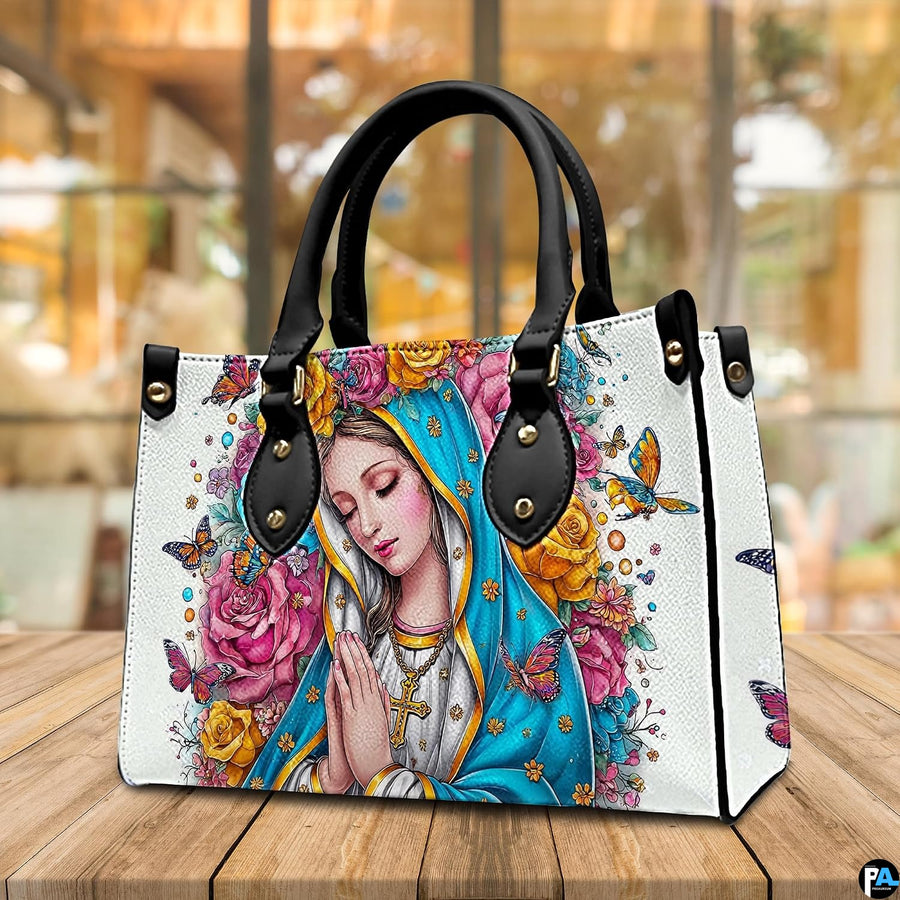 Praying Virgin Mary handbag displayed on a wooden tabletop in front of a sunlit window.