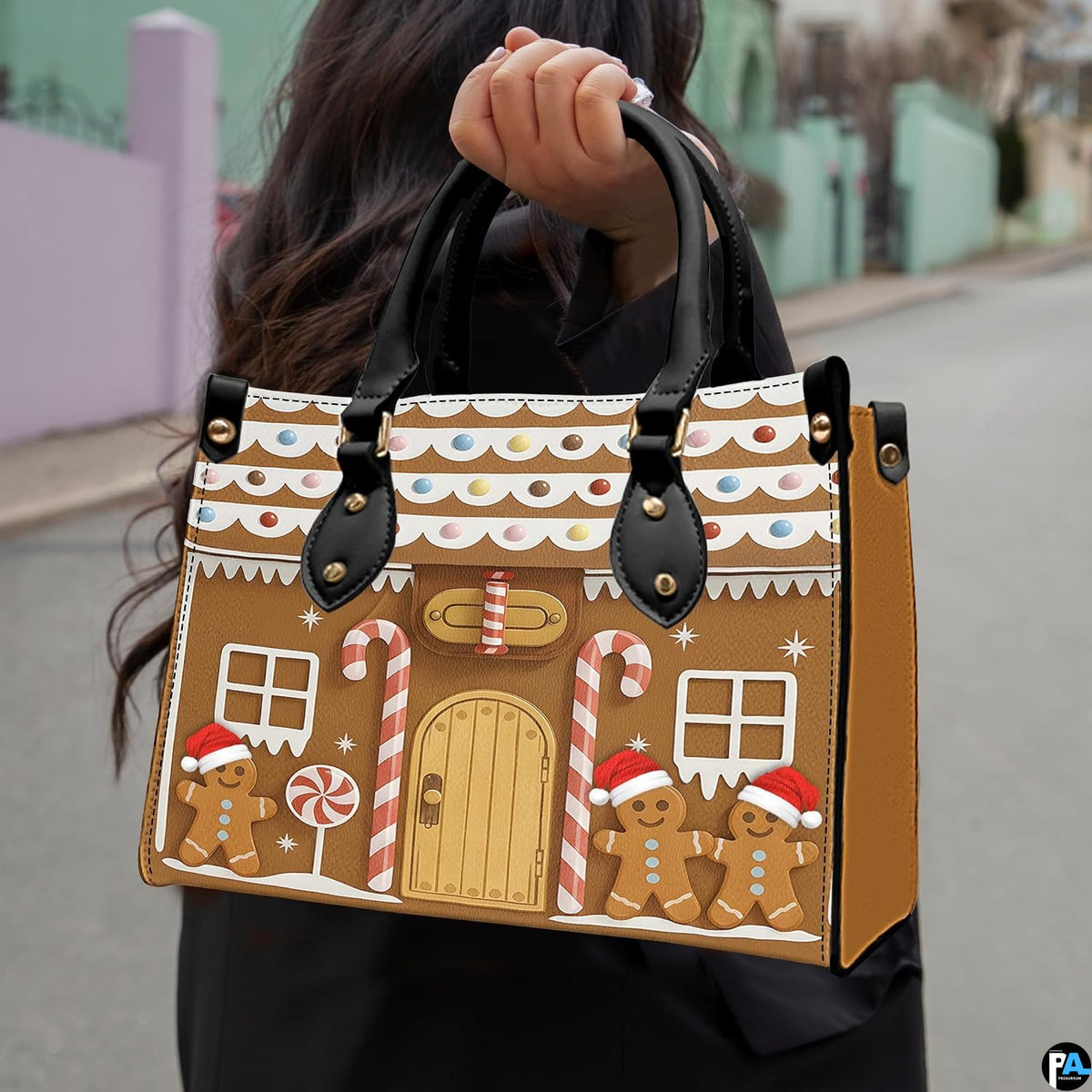 Model holding gingerbread house handbag by black top handles outdoors, displaying scale and silhouette.