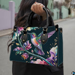 Model holding midnight teal handbag outdoors displaying flying hummingbird and botanical floral artwork.