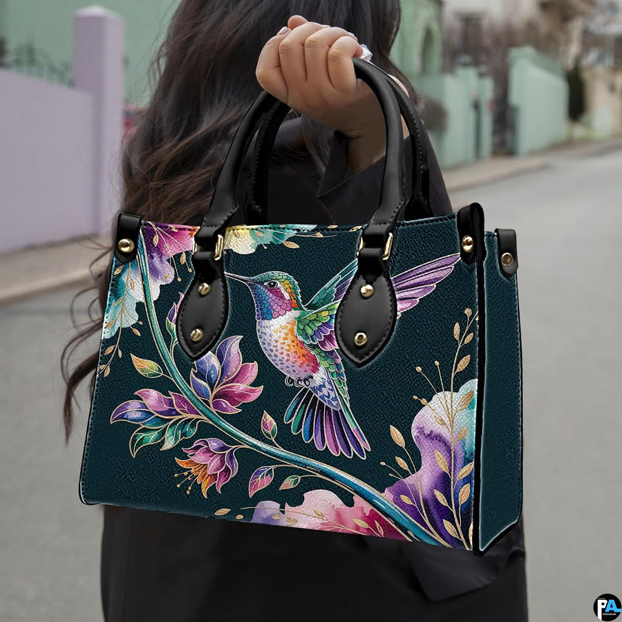 Model holding midnight teal handbag outdoors displaying flying hummingbird and botanical floral artwork.